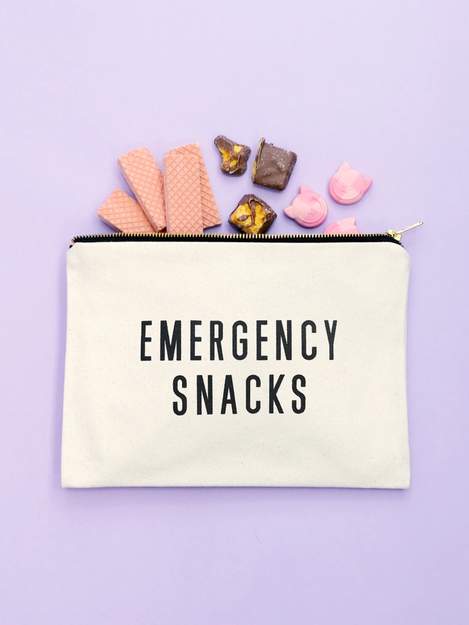 a natural canvas zip pouch with the words emergency snacks with biscuits and sweets spilling out on a purple backdrop