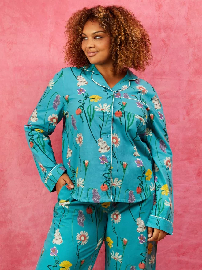 Model stands smiling off to the left with her hand in the pocket of a pair of traditional pyjamas with a classic shirt style top and wide legged bottom in a teal based print covered in wild british flowers in pops of pink, purple and yellow