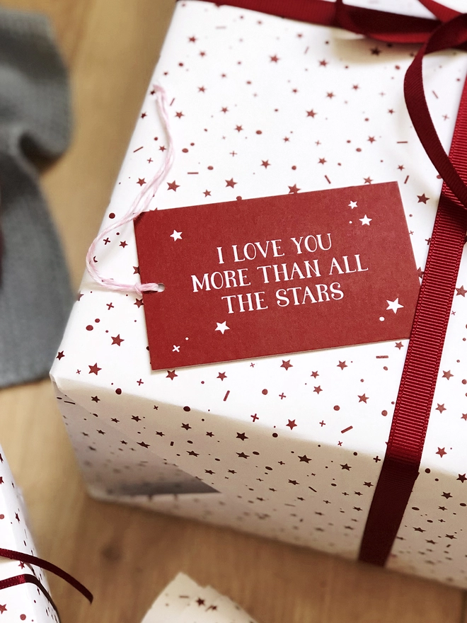 A gift wrapped in white wrapping paper with a red star design, tied with red ribbon, and a tag that reads "I love you more than all the stars" is on a wooden surface.