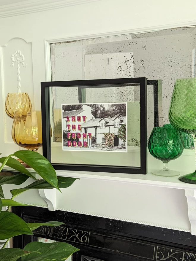Print of Shut the front door embroidered on B&W cottage in frame on shelf