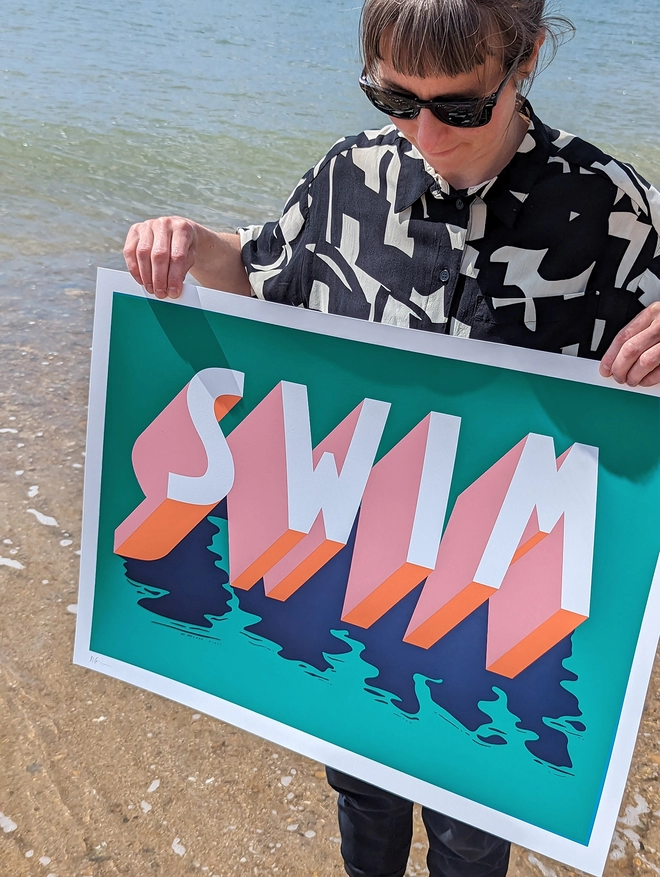 Artist Survival Techniques holding an art screenprint of the word SWIM in 3d typography in green, pink, orange and blue, standing in the sea at the beach.