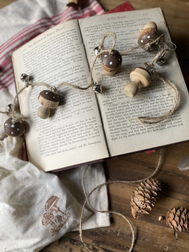 A string of Hand Painted Wooden Toadstool Bell Garlands trailing across an open book, on display with a small white pouch and some decorative pine cones