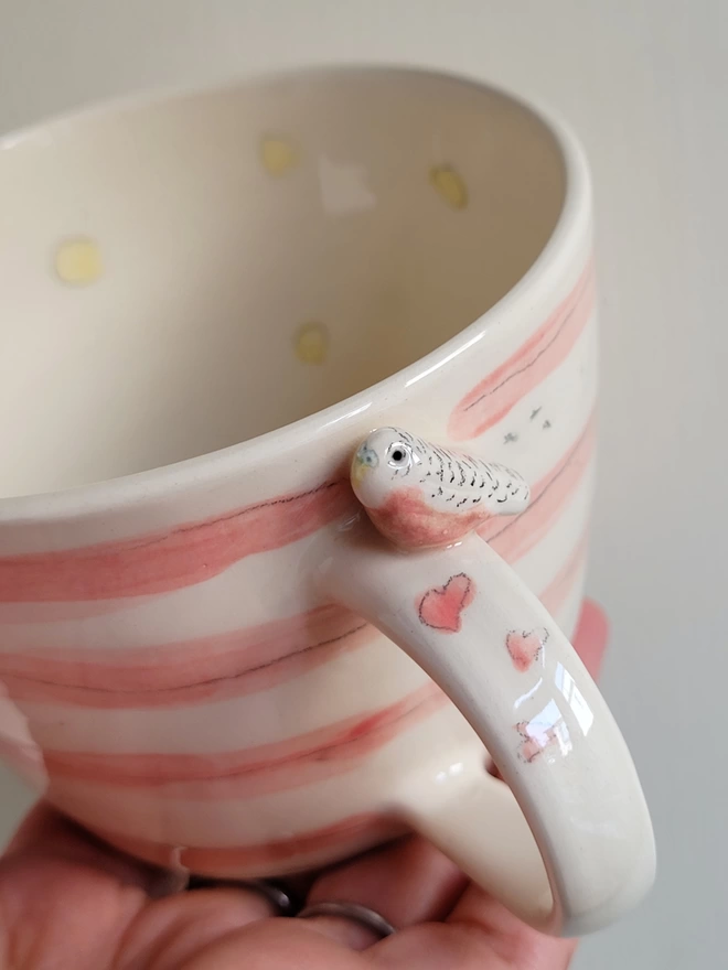 Kitsch quirky ceramic cup held in a hand it has a pink budgie end  hearts on the handle 