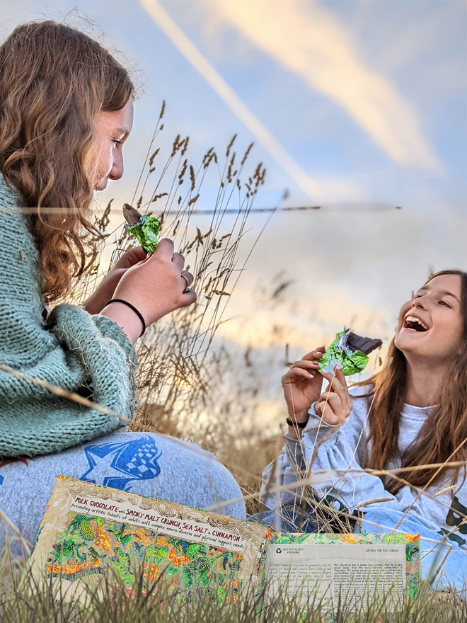 Two happy girls in a field enjoying charity milk chocolate wrapped in green foil & dino packaging
