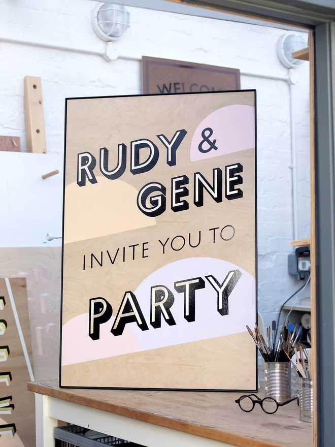 Handpainted invite to party sign with gold leaf letters outlined in black, with pastel coloured semicircles in the background. Shown in the artist's workshop. 