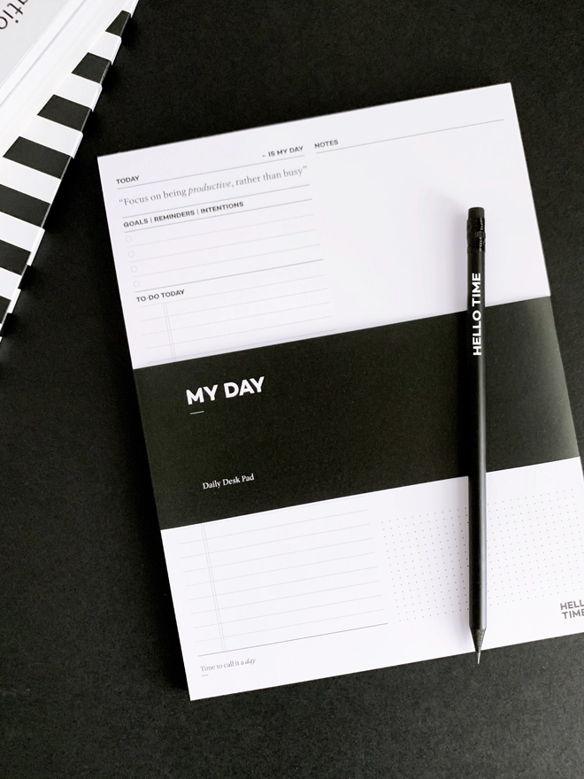 A MY DAY pad with a black HELLO TIME pencil laid on it on a black background.