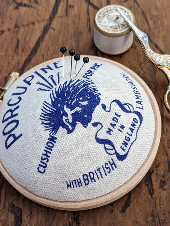 close up of royal blue porcupine embroidery hoop pin cushion shown with pins, scissors and cotton reel