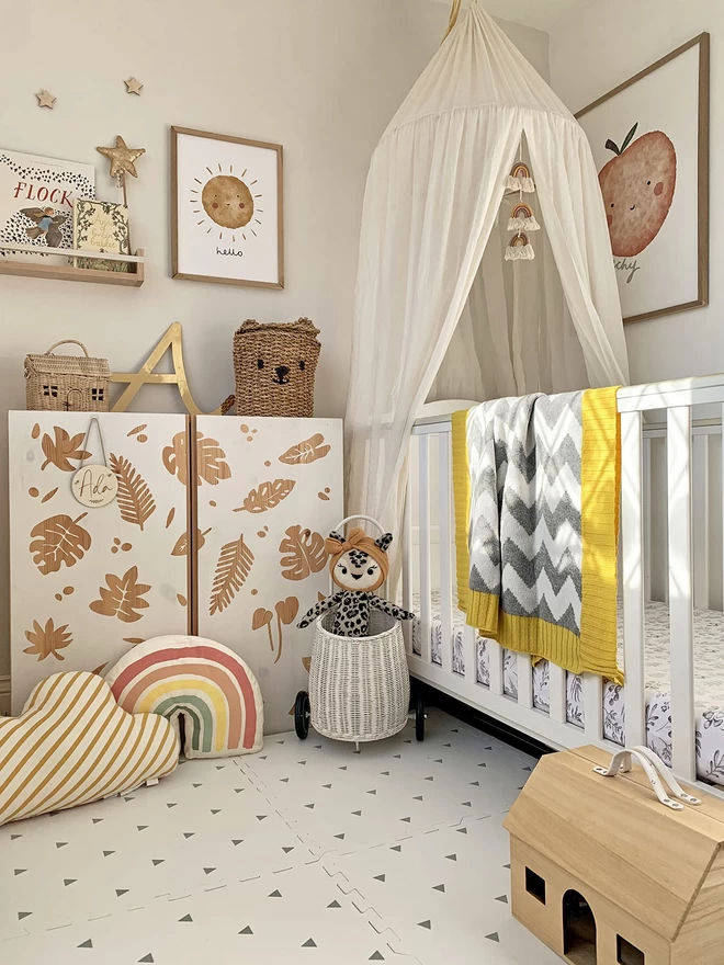 A neutral scandi style nursery with lots of natural wood, wicker accessories and a white cot. Over the cot is draped a grey and white chevron blanket with a mustard yellow trim. A poster on the wall features a cute poster saying ‘peachy’.