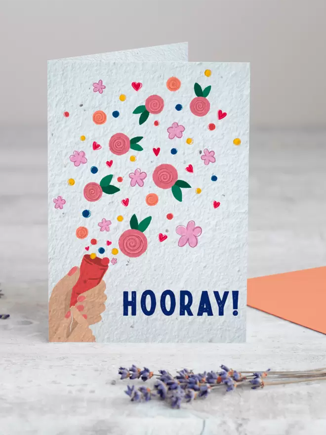 Plantable Card with ‘Hooray’ in the bottom right of the card with an illustrated hand pulling a party popper with confetti and flowers coming out with a sprig of Lavender in the foreground
