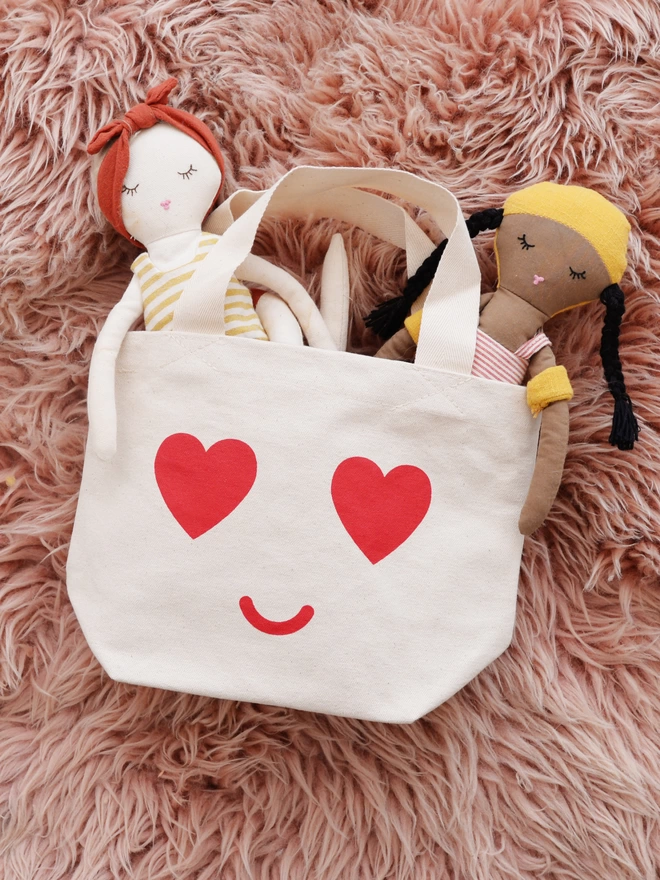 a mini kid's size tote bag with heart eyes and smiling mouth laying on a pink fluffy rug with two dolls sticking out