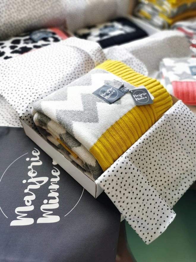 A neatly folded chevron blanket is shown packed inside a box and wrapped with tissue paper. The box is sitting on a table surrounded by other gift wrapped products. Beneath the box is a grey Marjorie Minnie branded gift bag.