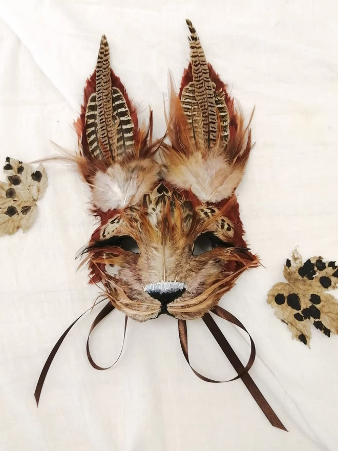 A luxury brown hare masquerade mask laying flat next to dried leaves