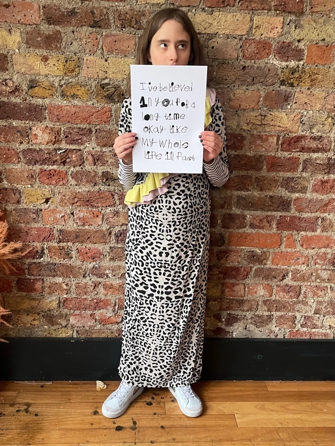 full length picture of Piper a teenage deaf, autistic, teenage artist from hackney, wearing a full length leopard print dress holding her artwork which is black handwritten unique font on a white background.