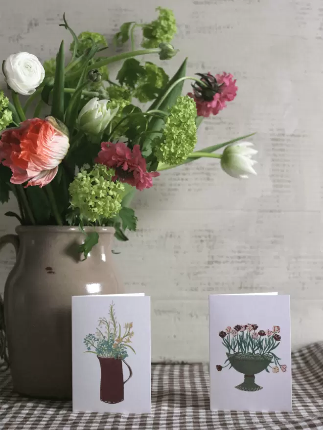 2 flower cards on a gingham tablecloth next to a bunch of fresh flowers in a beige vase. 