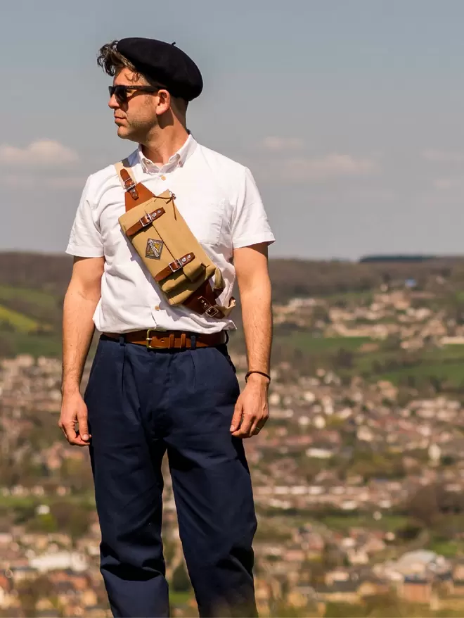 Model wearing taupe sling bag with leather trim standing with village in the background.