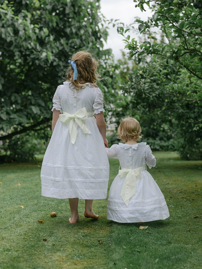Two girls in white dresses with yellow sashes walk away from the camera  