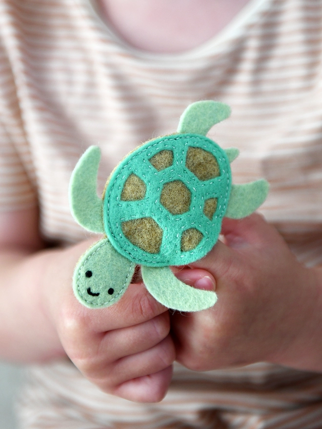 A young child holds a green felt sea turtle finger puppet on their fingers. 