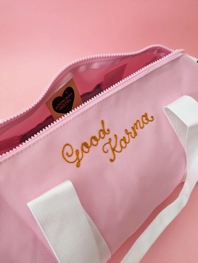 Pink Duffel Bag close up detail with white straps and handles, personalised with gold embroidery reading 'Good Karma' on a pink background showing the bag open