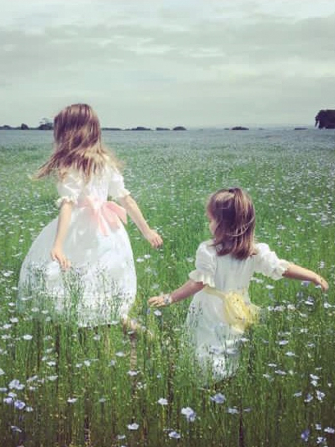 Two girls walk through a field of flowers in white dresses with sashes