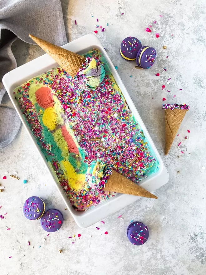 a bowl of colourful unicorn ice cream with colourful sprinkles on top and bright purple macarons