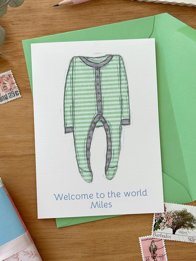 An illustrated new baby greetings card with a green onesie design lays on a green envelope on a wooden desk.