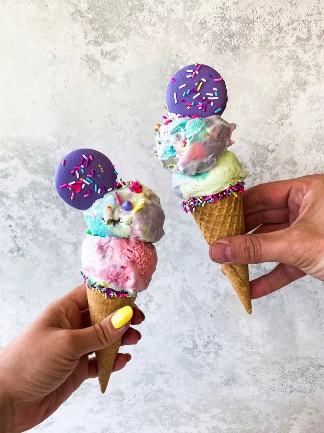 two hands holding two ice cream cones decorated with a chocolate rim and colourful sprinkles, with colourful scoops of ice cream with sprinkles & a purple macaron with sprinkles on top of each ice cream
