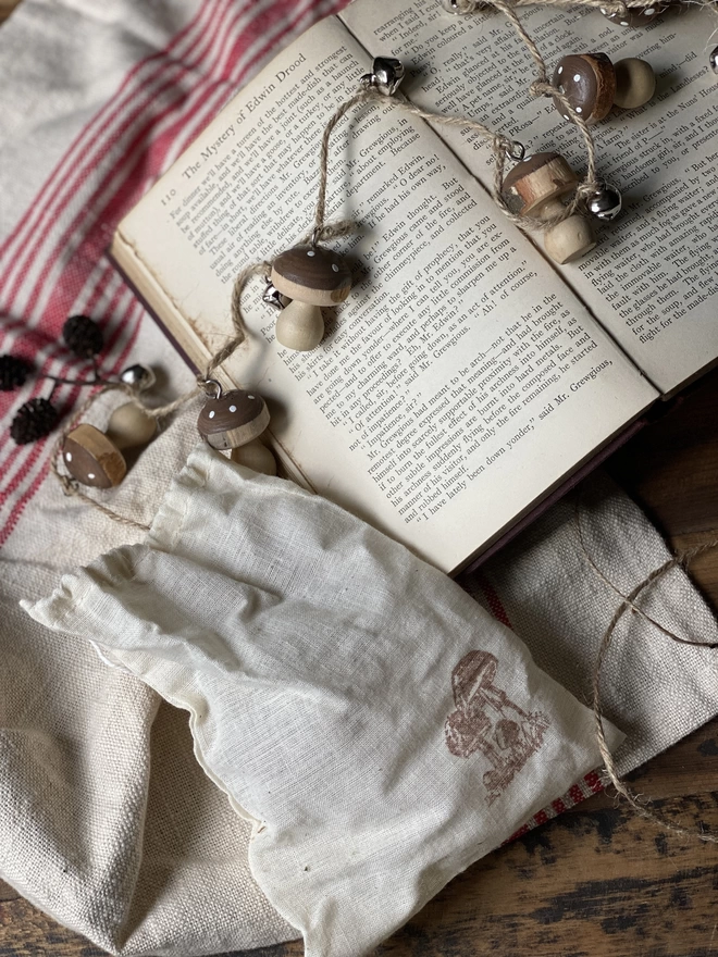 A string of Hand Painted Wooden Toadstool Bell Garlands trailing across an opened book atop a red and white striped cloth, with a small white pouch in front