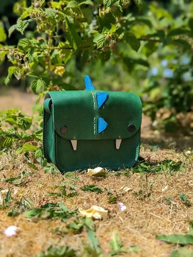  Handmade 'Bagasaurus' cross- body handbag in hand- dyed green leather with forward- facing turquoise spikes., pictured against a garden backdrop.