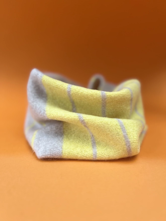 Yellow oatmeal knitted striped snood shown from the side on an orange background