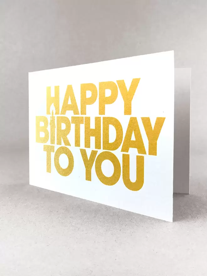 Happy Birthday to You landscape greetings card. The I of Birthday is a candle using negative space. A ¾ view to show the plain inner. Printed in capital letters, simple font printed in a gold ink on a white card made by Salty's Studio.