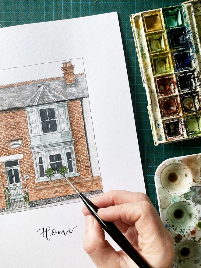 A white page inside and a hand painting a beautiful Edwardian style house in the centre painted in intricate watercolour details. Below black calligraphy lettering reads home. The background is a green cutting mat and the paint palette which is well used can be seen to the right.