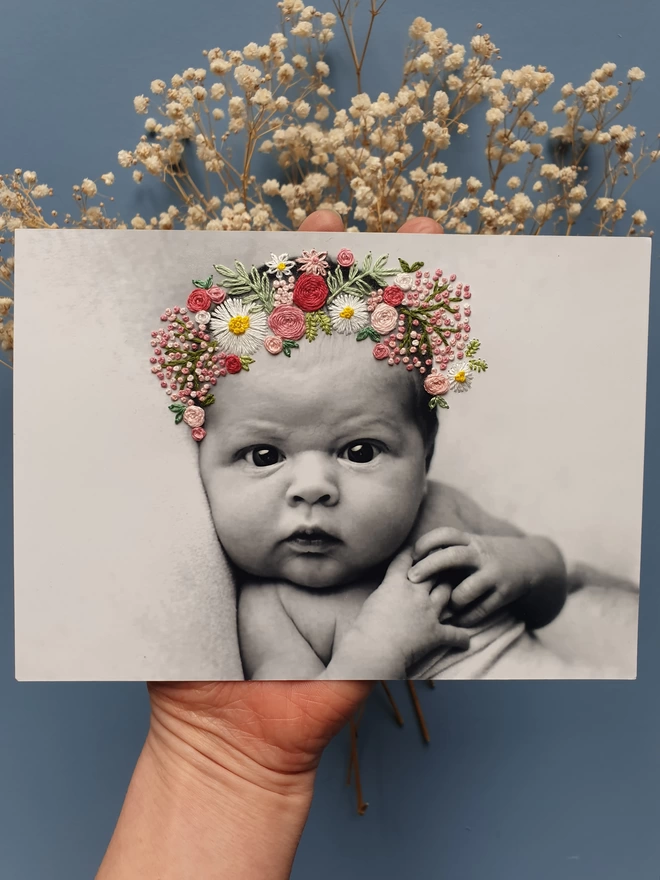 Baby photo in B&W with hand embroidered pinks and white flower crown, held over blue background