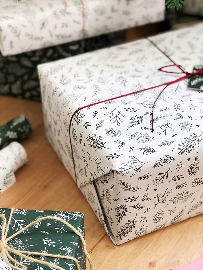 A gift wrapped in white wrapping paper with a Christmas botanical design lays on a wooden floor.