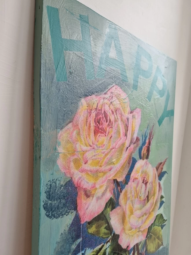 A painted plywood panel in turquoise and blue featuring decoupage of large pink roses taken from a Victorian seed packet with a motivational phrase that ‘happy’.