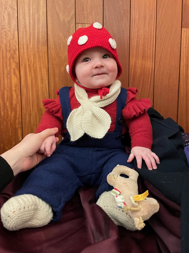 A baby in a variety of knits wears a red crocheted bonnet with white dots