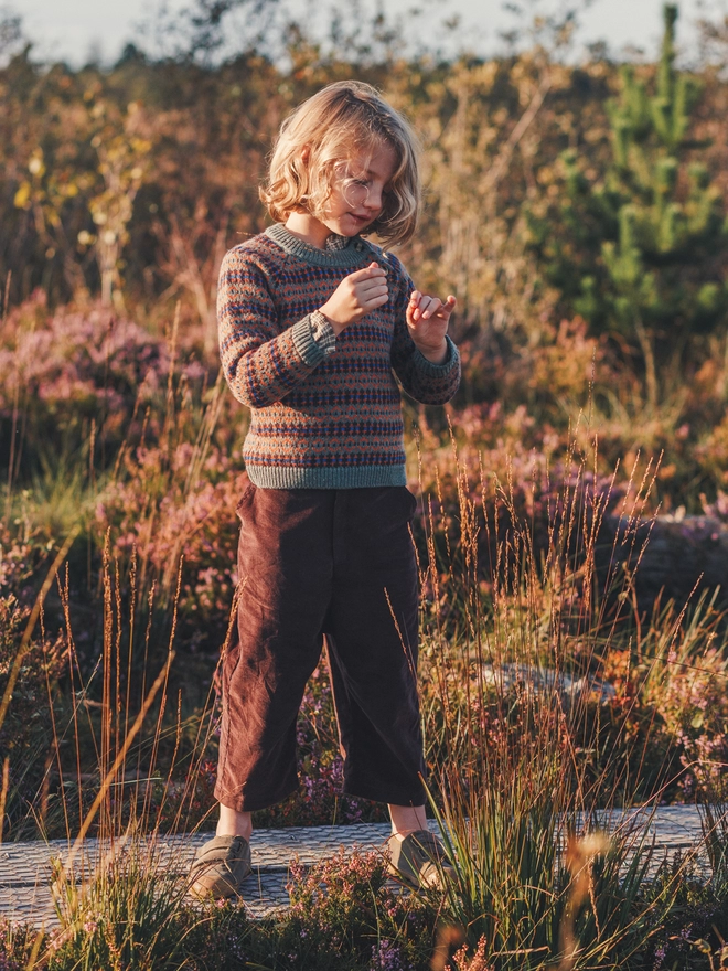 A boy stands in the middle of a bog strewn in heather wearing a moss green fairisle jumper and brown trousers