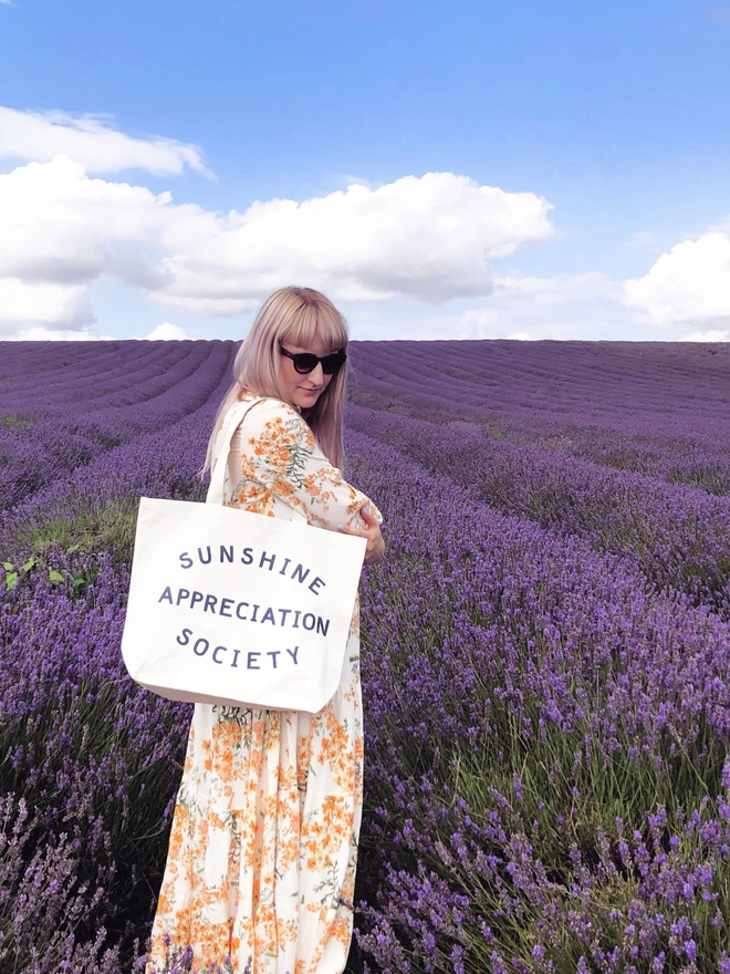 a model standing in a lavender field carrying a large natural canvas tote bag bearing the words sunshine appreciation society 