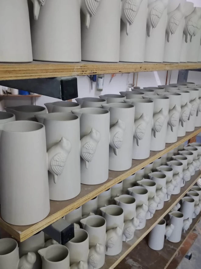 Handmade clay bird jugs are neatly lined up on shelves, ready to go in the kiln for a bisque firing. 
