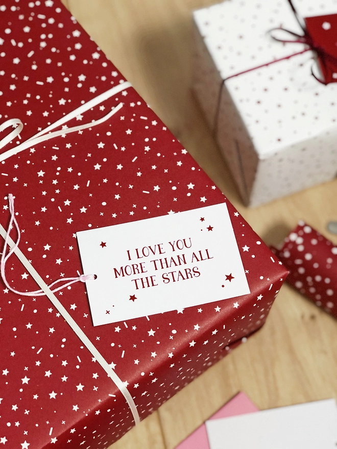 A gift wrapped in a red star design wrapping paper and a tag that reads "I love you more than all the stars" is on a wooden surface.