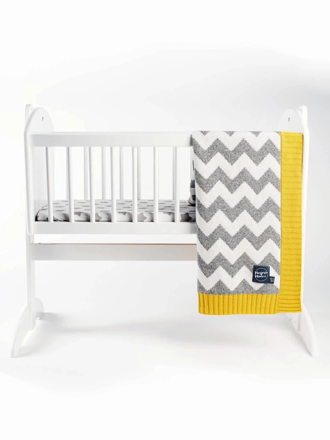 A product shot of a white rocking crib against a white background. A grey and white zigzag pattern baby blanket with mustard yellow ribbed trim is draped over the side of the crib.