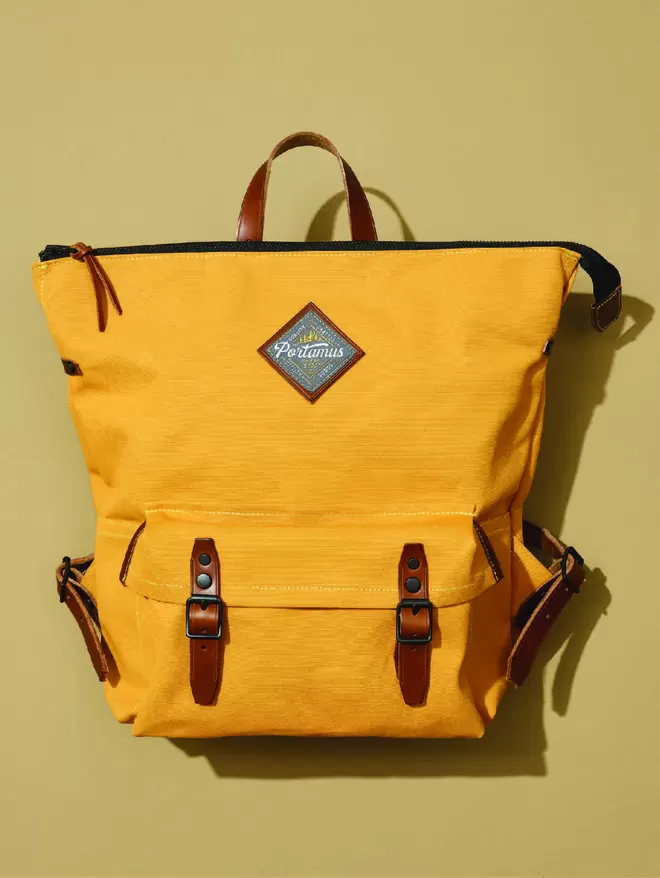 Yellow zip top backpack with brown leather trim and black hardware on taupe background.