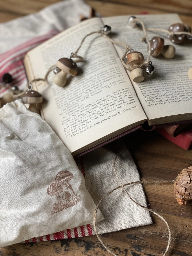 A string of Hand Painted Wooden Toadstool Bell Garlands trailing across an opened book atop a red and white striped cloth, on display with a small white pouch