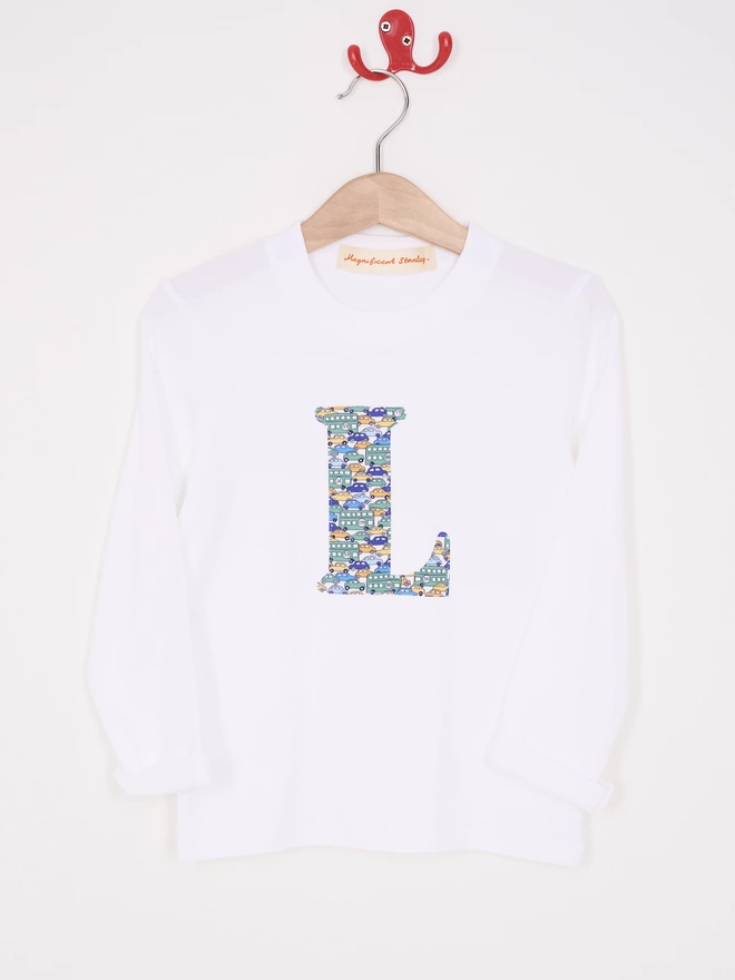 A white cotton long sleeve t-shirt appliquéd with an initial in a Liberty print featuring cars and busses, hung on a hanger 