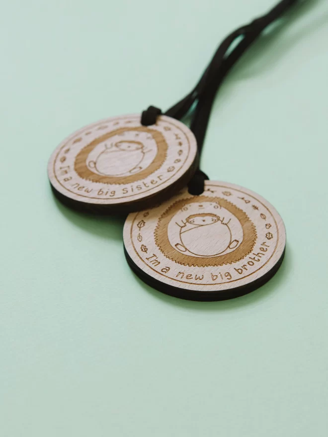 2 wooden medals for new big siblings. The medals are etched with a drawing of a hedgehog holding a baby hedgehog