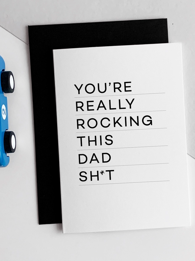 Rocking Dad Birthday Card that reads "You're really rocking this dad sh*t" overlapping a black envelope beside a blue racing car toy.