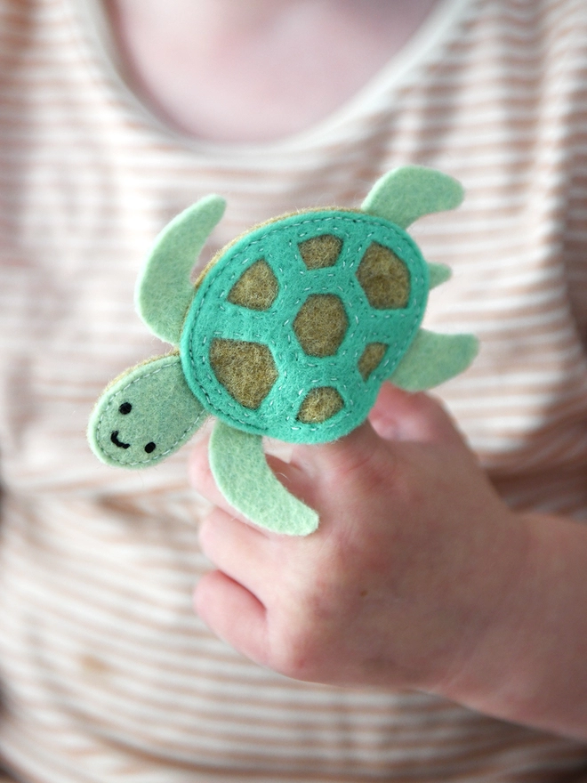 A young child holds a green felt sea turtle finger puppet on their fingers. 