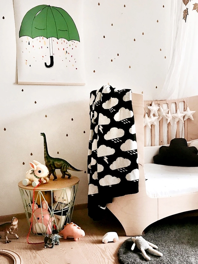 A neutral scandi style nursery showing a rounded wooden crib with a black and white storm cloud junior blanket draped over the side. Above the cot hangs a print of a green umbrella crying rainbow tears. There is metal basket on the floor containing soft toys that are spilling out onto the floor.