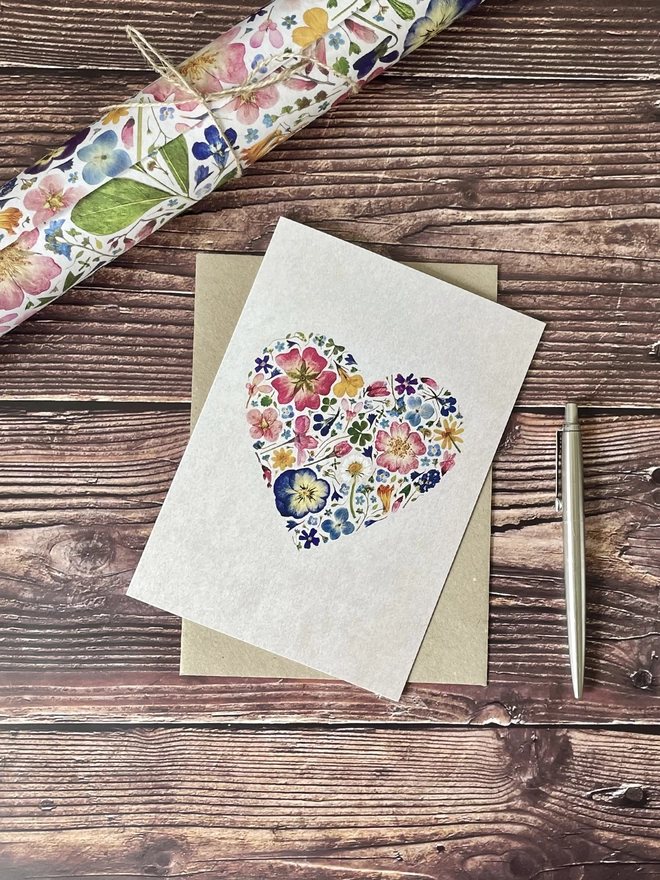 Floral Greetings Card with Pressed Flower Heart Design and Brown Kraft Envelope - Dark Wooden Background - Silver Parker Pen - Rolled Pressed Flower Wrapping Paper Tied with Brown String