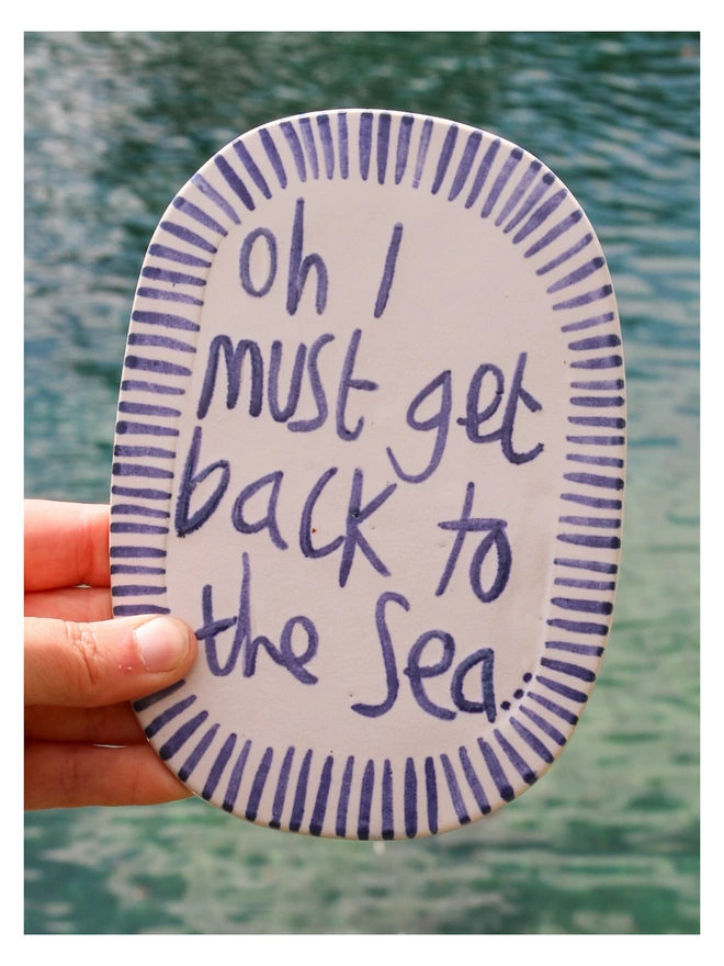 "oh i must get back to the sea" blue and white wall plaque with stripy edge in front of the sea