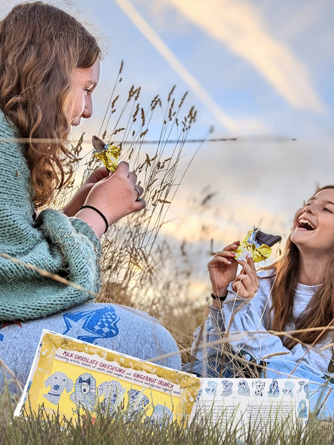 Two happy girls in a field enjoying charity milk chocolate wrapped in gold foil & blue dog packaging